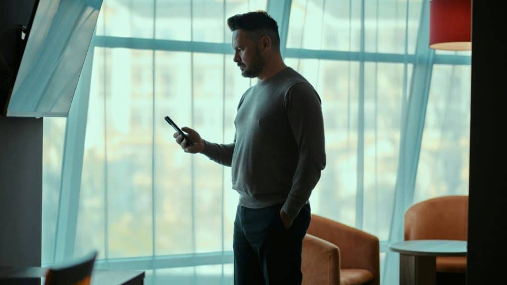 A bearded man stands by a window looking down at his mobile phone.