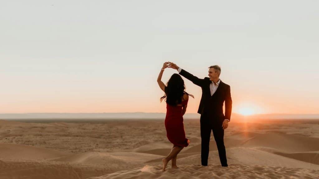A couple dancing together on sand dunes at sunset, silhouetted against the horizon.
