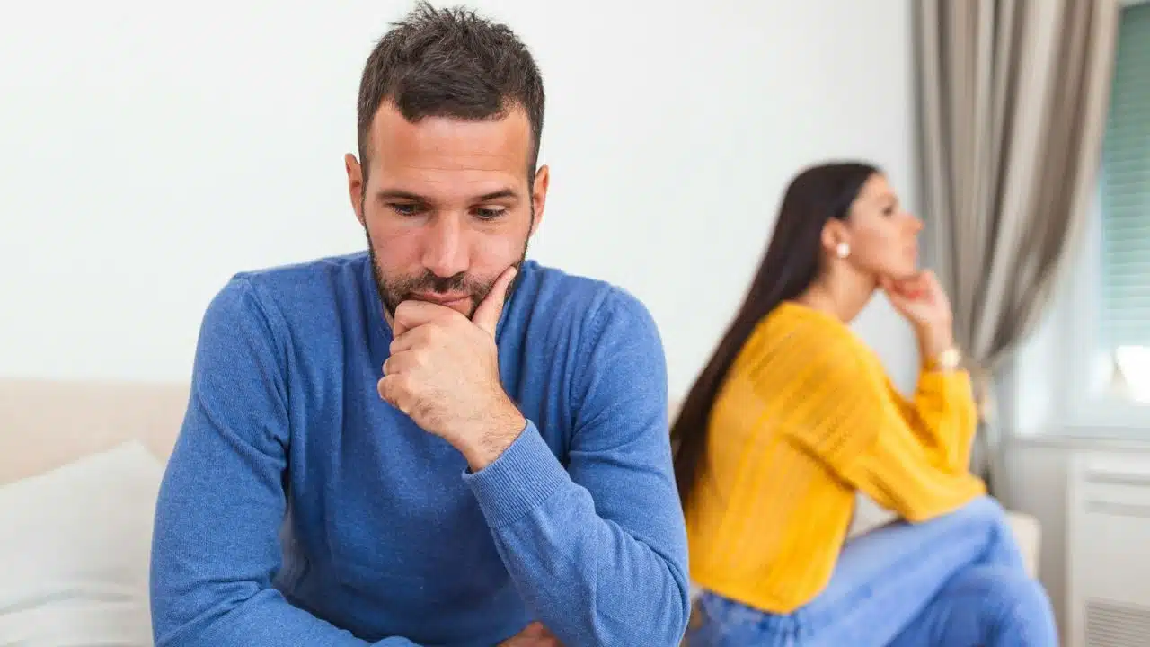 A man in a blue sweater looks down while a woman in yellow sits facing away.