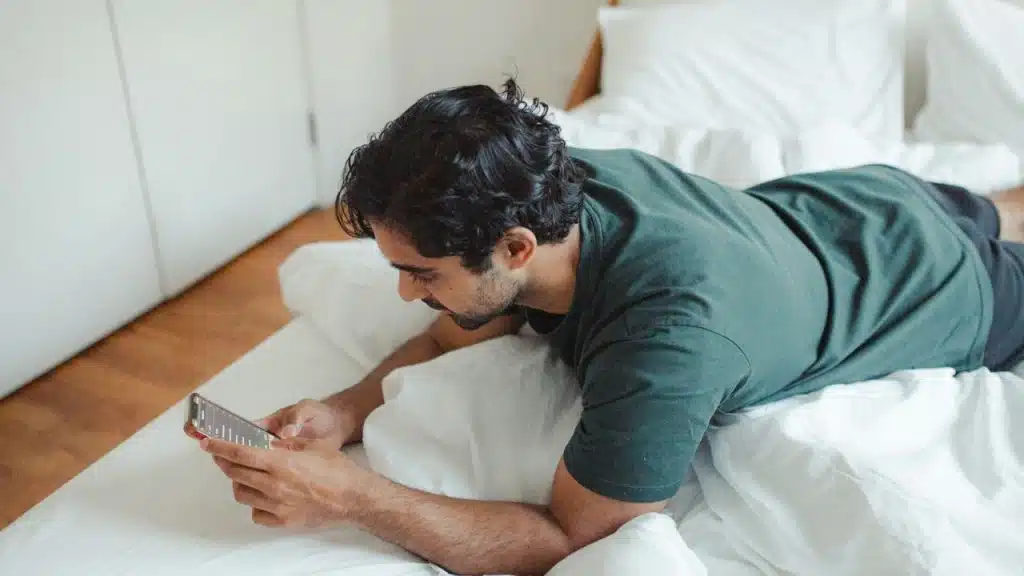 A man is lying on his stomach in bed, looking at his smartphone.