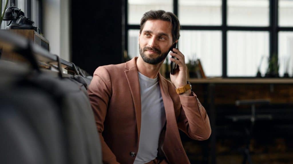 A bearded man in a brown blazer smiles while talking on a smartphone indoors.