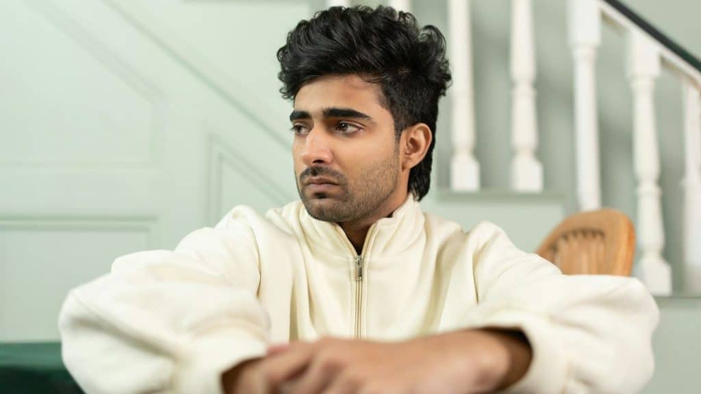 A man with dark, styled hair and a cream-colored jacket gazes thoughtfully to the side.