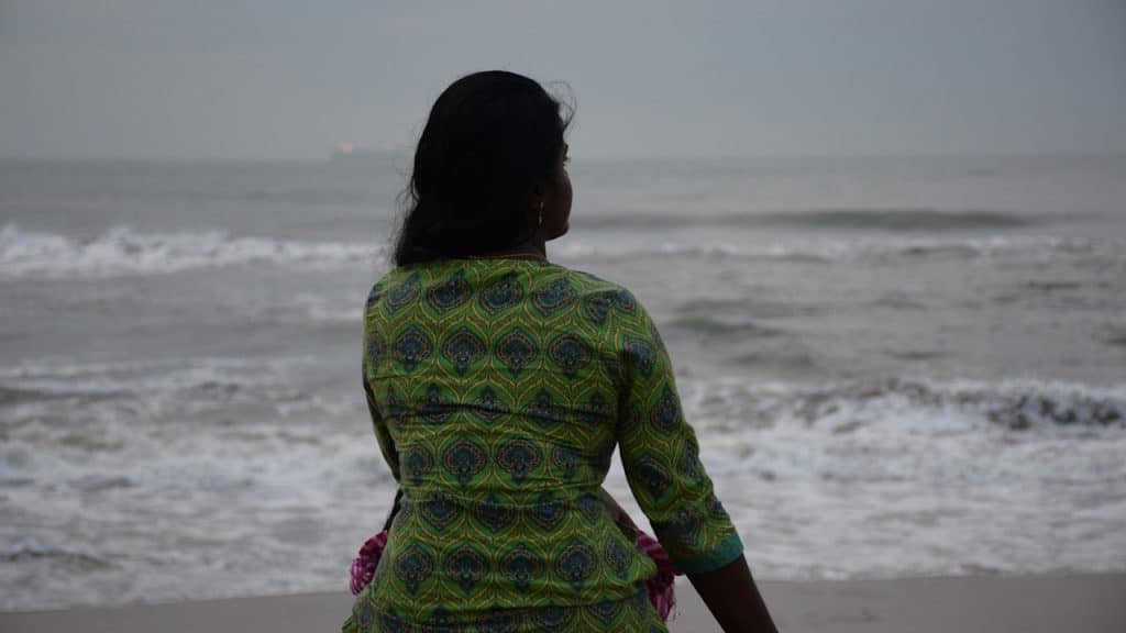 A person seen from behind, wearing a green patterned top, sitting on a sandy beach and looking out at the ocean waves under an overcast sky.