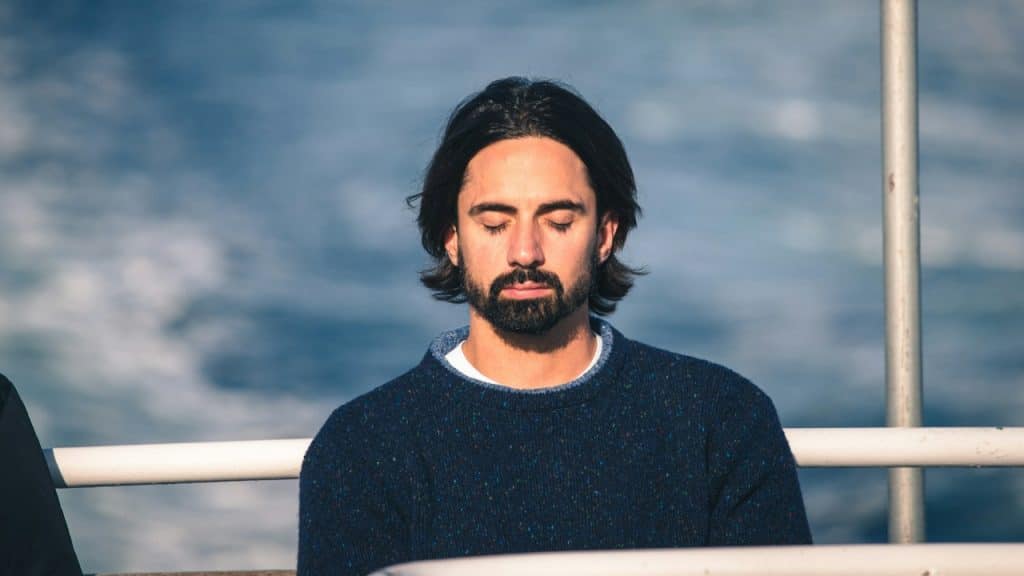 A man with a beard and closed eyes meditates outdoors with water in the background.