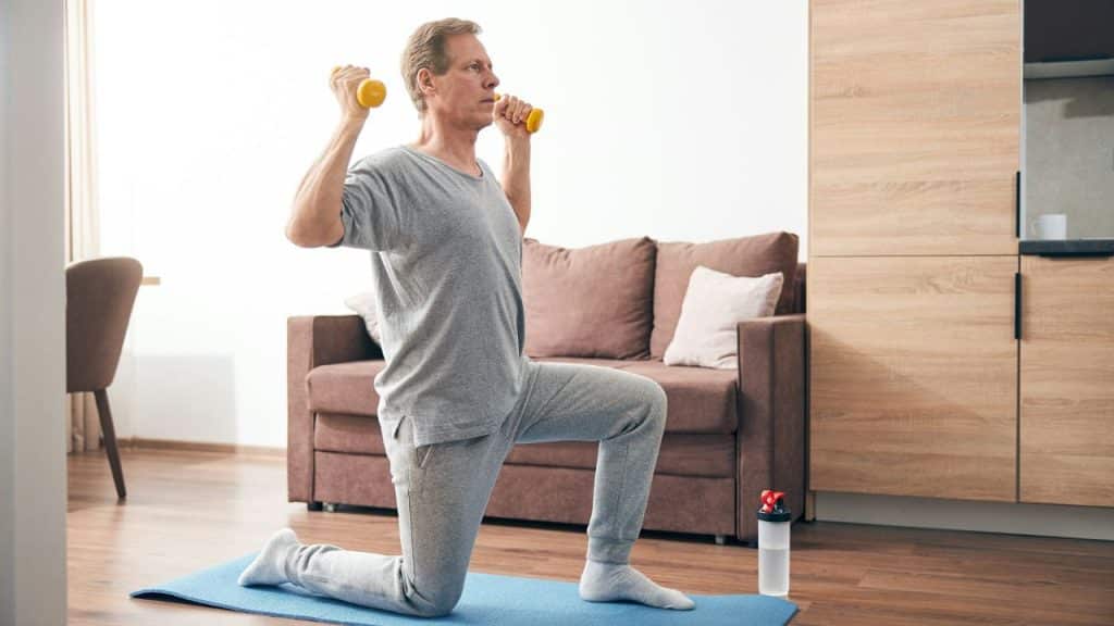 A man in a living room kneeling on a blue mat while holding small yellow dumbbells.
