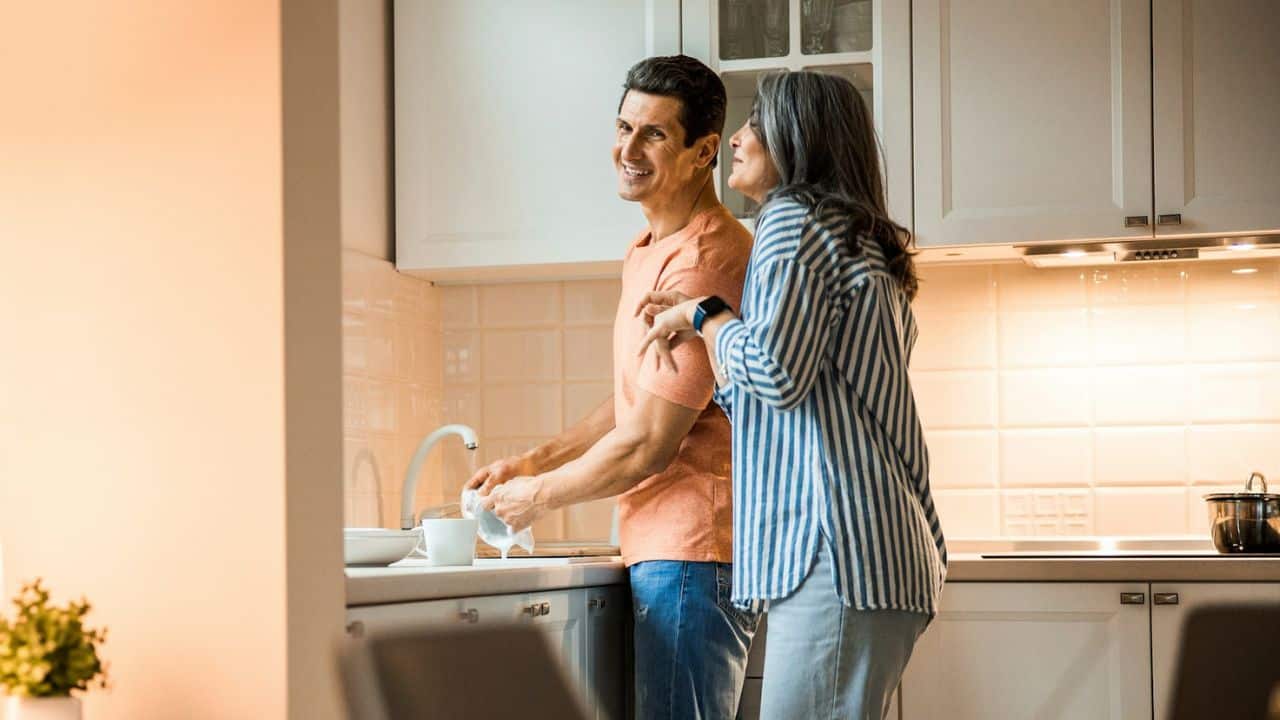 A man washes dishes in a kitchen while a woman stands beside him and smiles.