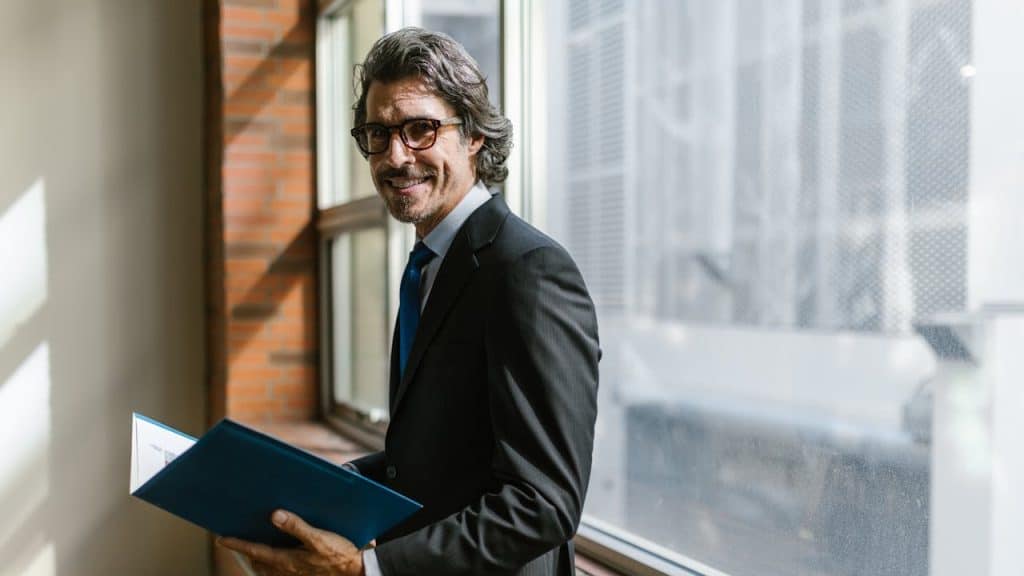 A smiling man in a business suit holding a blue folder while standing near a window in an office setting.