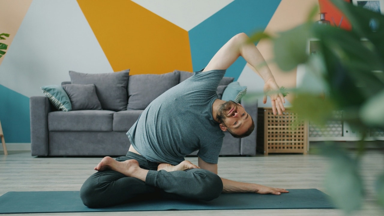 A man in a grey t-shirt performs a seated side stretch on a yoga mat.