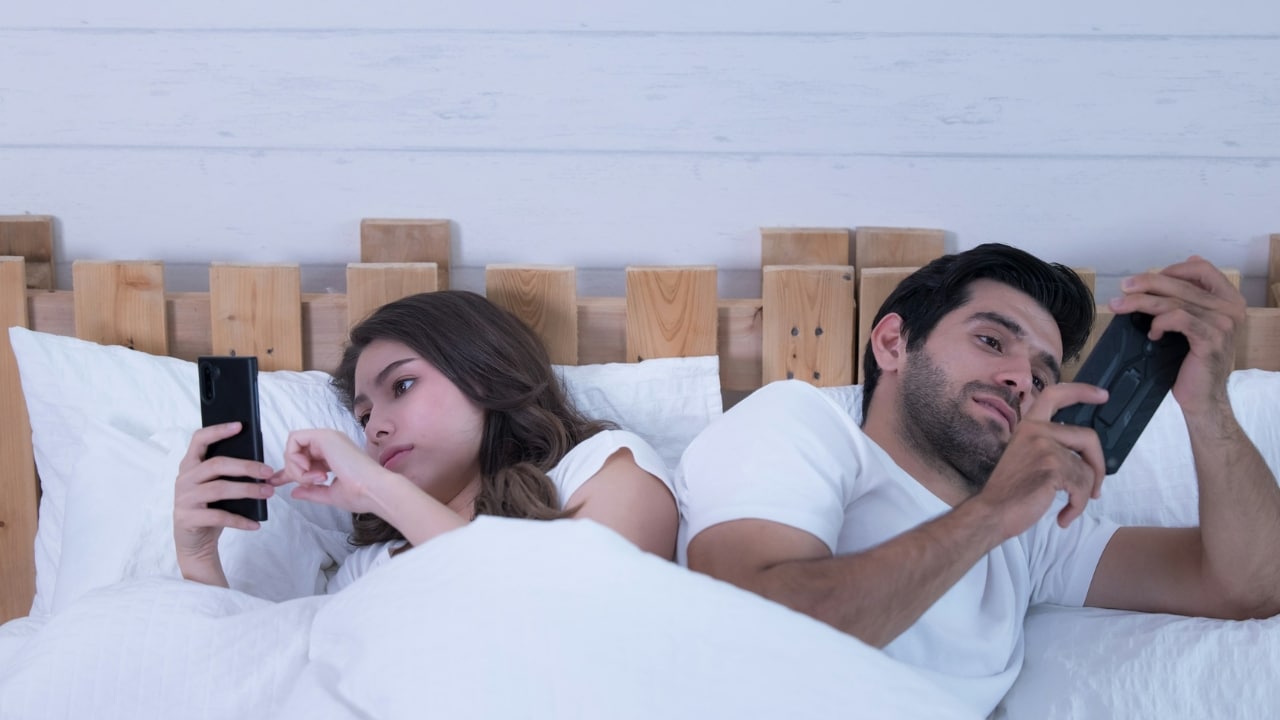 A man and woman lie in bed back-to-back, each looking at their own smartphone.