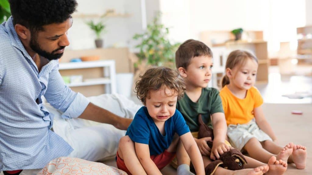 A man sits on the floor near three children, including one toddler who is crying.