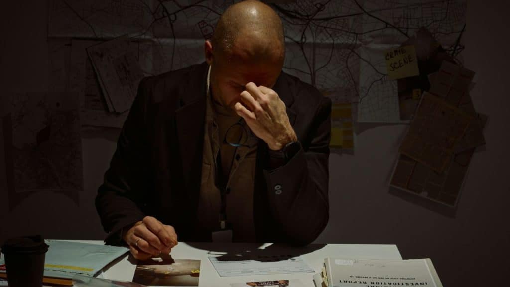 A bald man sits at a desk holding the bridge of his nose near documents.