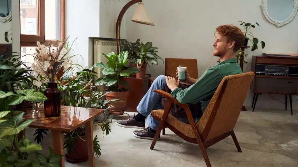 A man sits in a chair holding a mug in a room filled with potted plants.