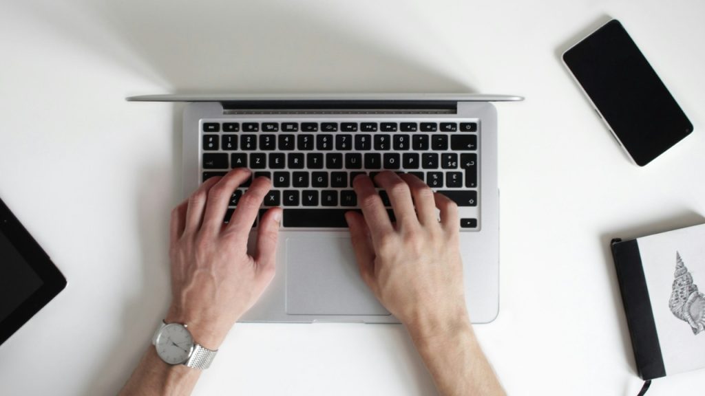 Overhead view of hands typing on a laptop surrounded by a phone and notebook.