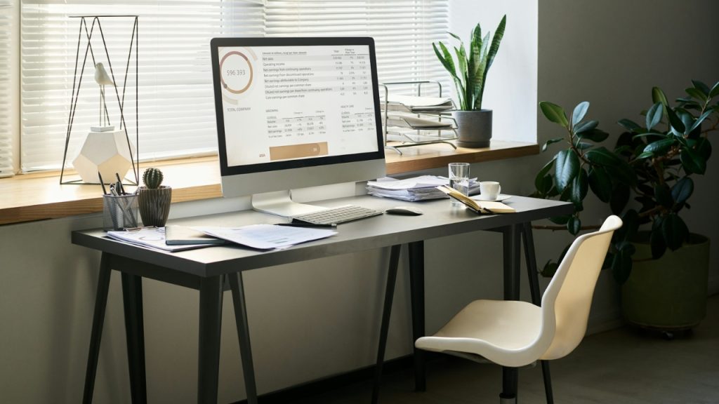 A clean desk with a computer monitor, plants, and papers sits in front of a window.