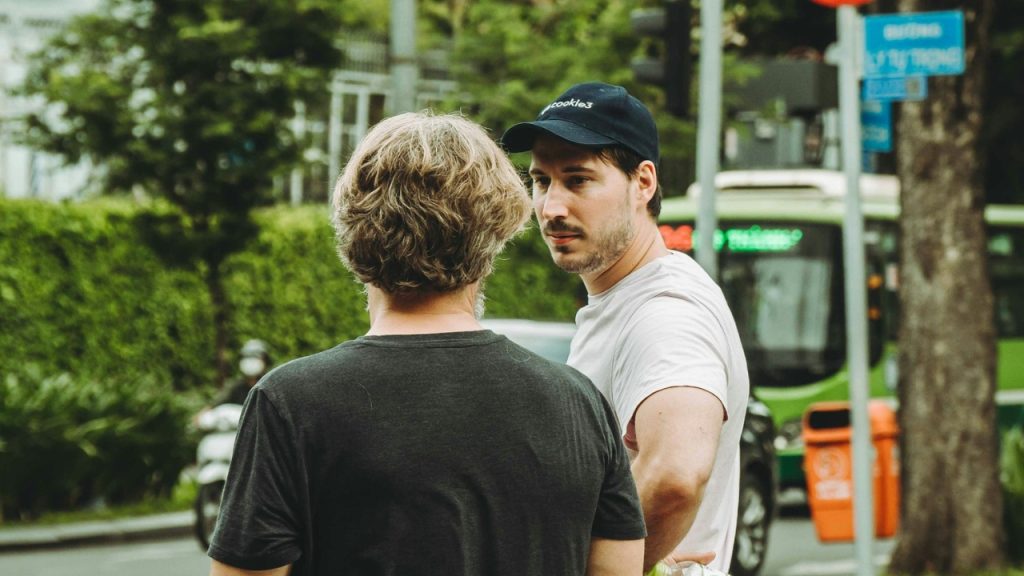 A man in a baseball cap looks at another man while standing near a city street.