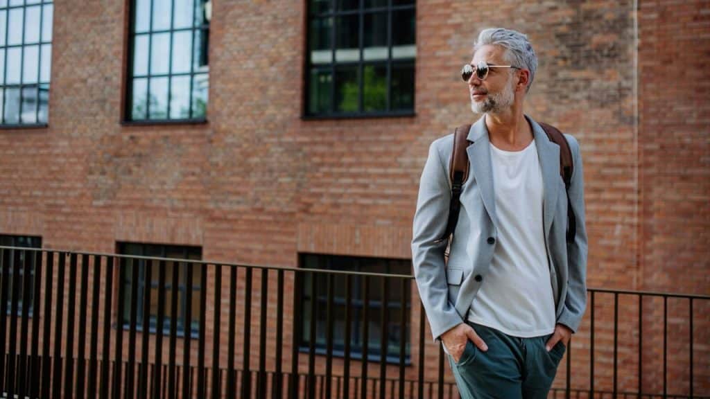 A grey-haired man in a blazer and sunglasses stands outdoors in front of a brick building.