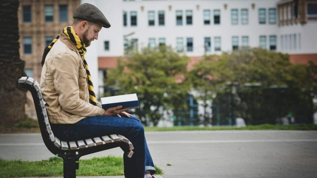 A man in a flat cap and yellow scarf sits on a park bench reading a book.