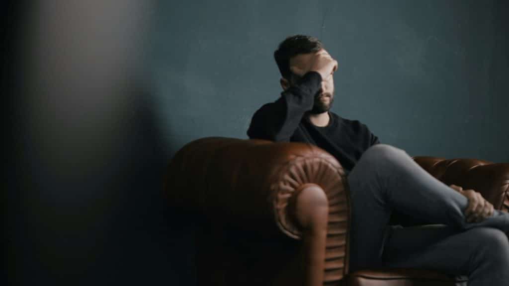 A man sits alone in a leather armchair with one hand covering his forehead.