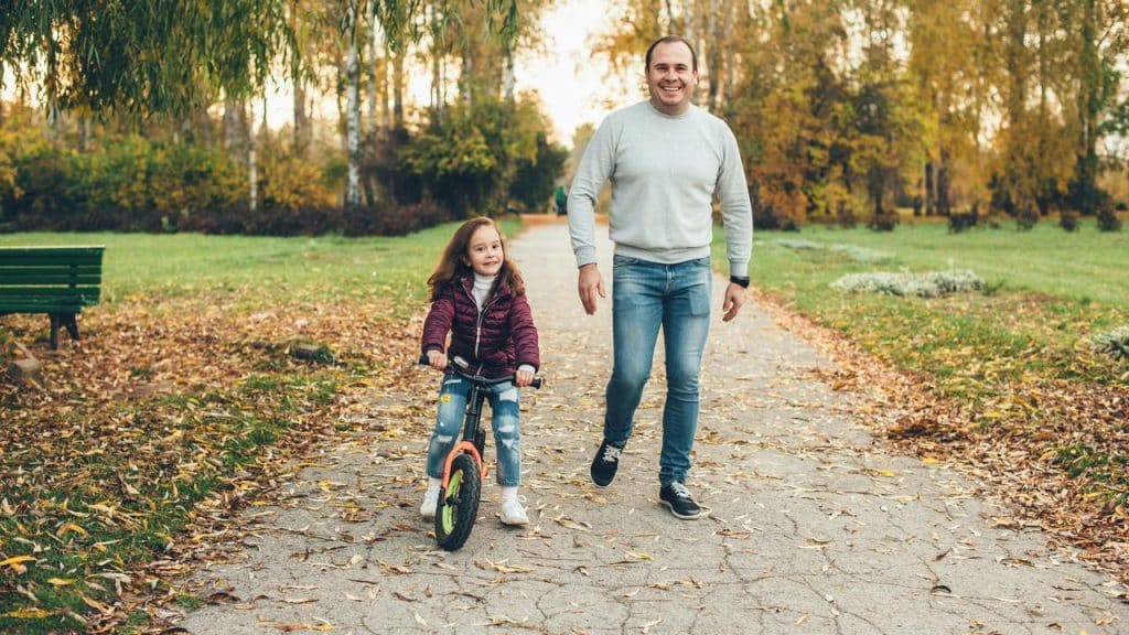 A man walking beside a girl riding a bike on a leafy park path.