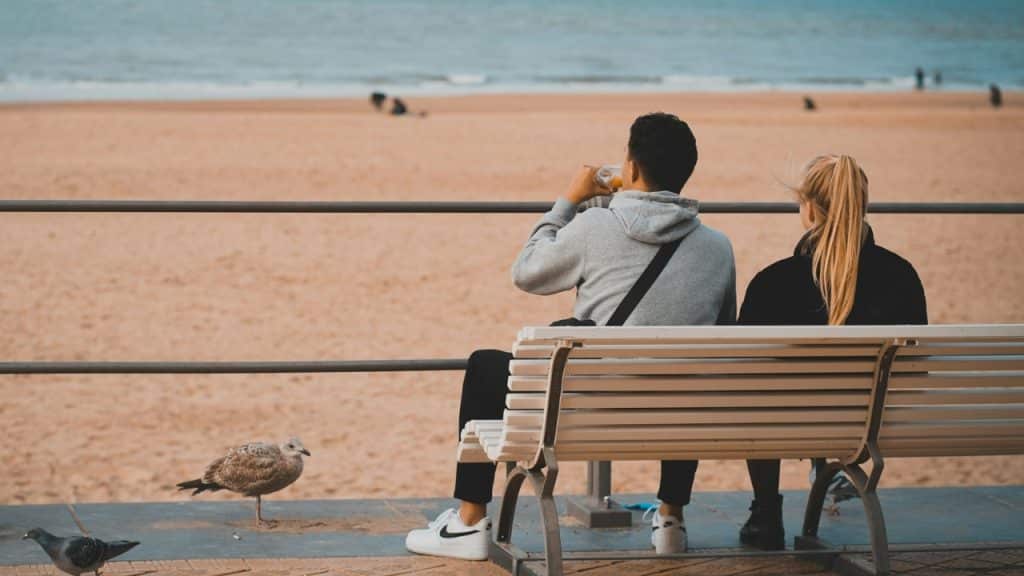 A man and woman sit on a bench facing a sandy beach with several birds nearby.