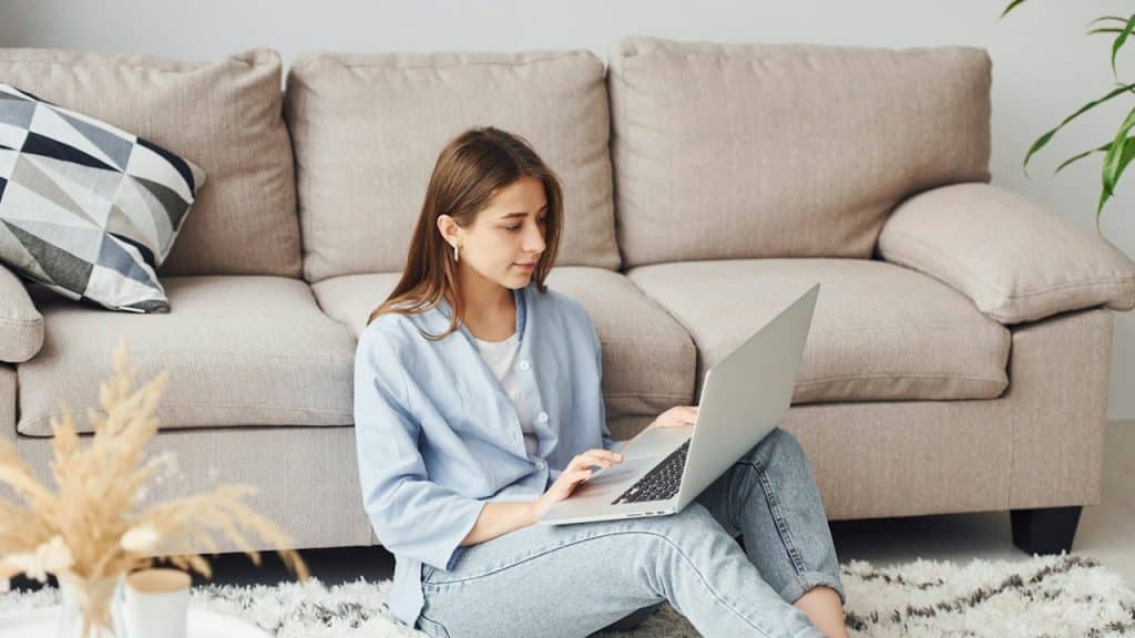 A beautiful-young-woman-in-jeans-and-blue-shirt-sits-on-the-floor-with-laptop-indoors-at-home.