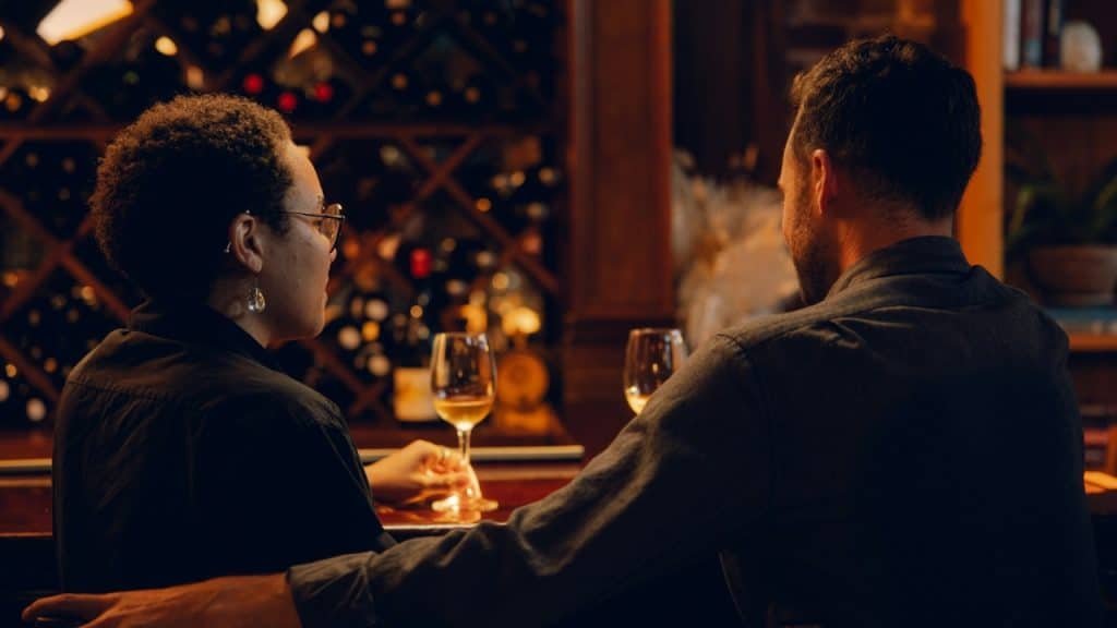A man and woman sit at a bar with wine glasses, seen from behind.