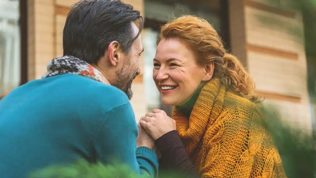 A man and woman hold hands and smile at each other while outdoors near buildings.