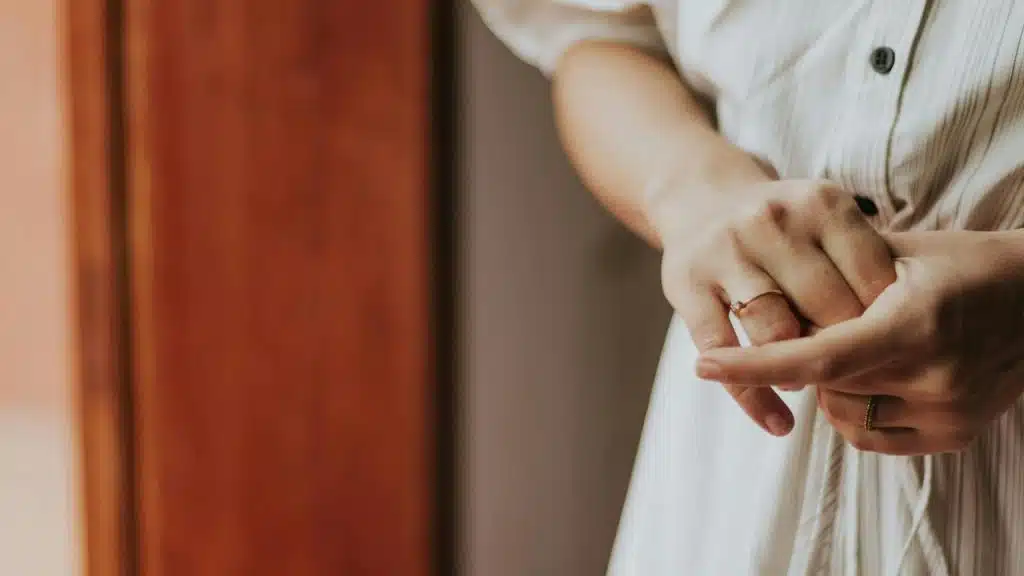 A person clasping their hands while wearing a light-colored dress indoors.