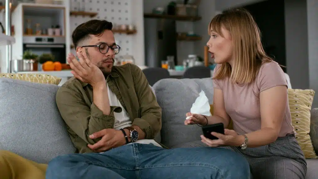 A woman speaks to a man while holding a phone and a tissue on a sofa.