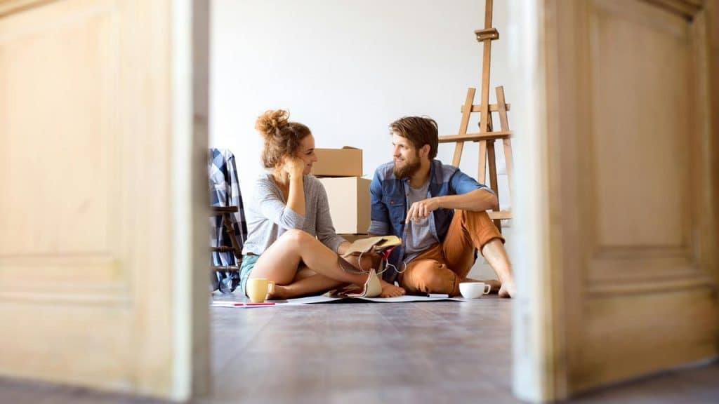 A couple sitting on the floor at home, talking and unpacking boxes.
