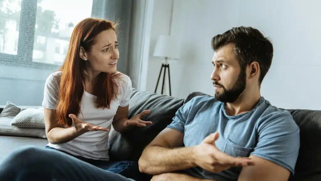 A woman arguing with a bearded man who gestures with his hand while they sit on a couch.