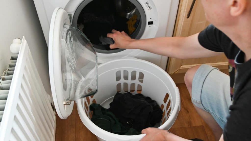 A person loads clothes into a front-loading washing machine beside a laundry basket.