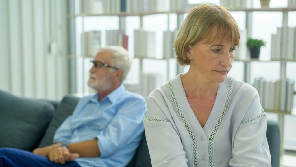 An older woman looks downward while an older man in a blue shirt sits behind her.