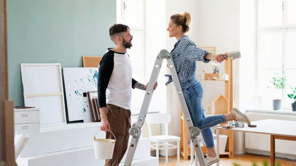 A man holds a paint bucket while a woman stands on a ladder holding brushes.