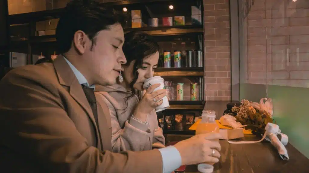 A man in a suit speaks while a woman beside him drinks from a paper cup.
