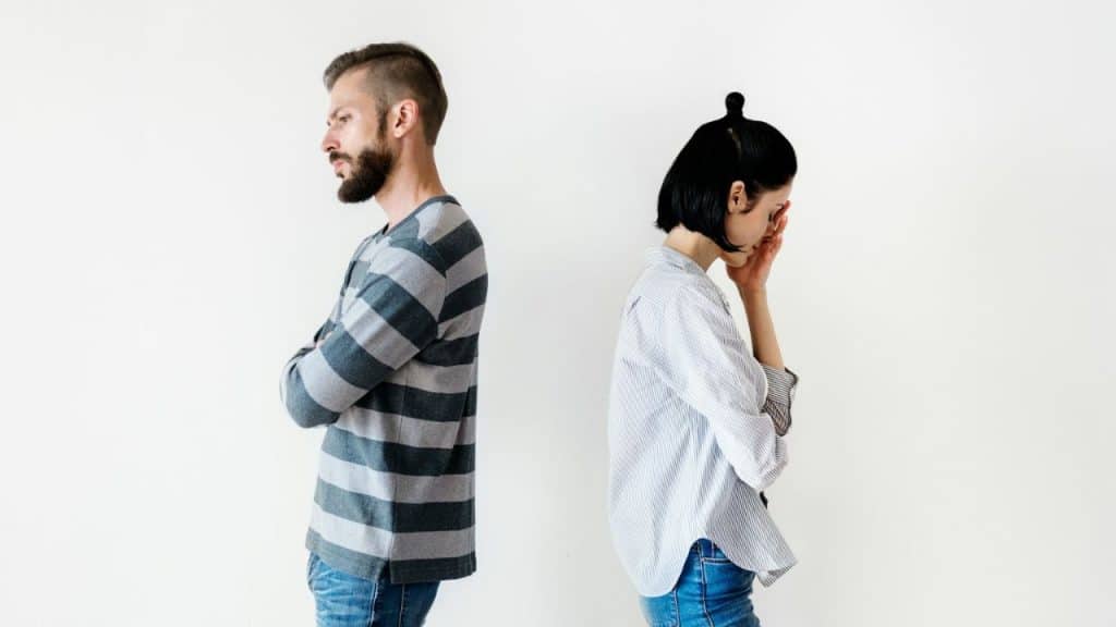 A man with a beard and a woman stand back-to-back against a plain white wall.
