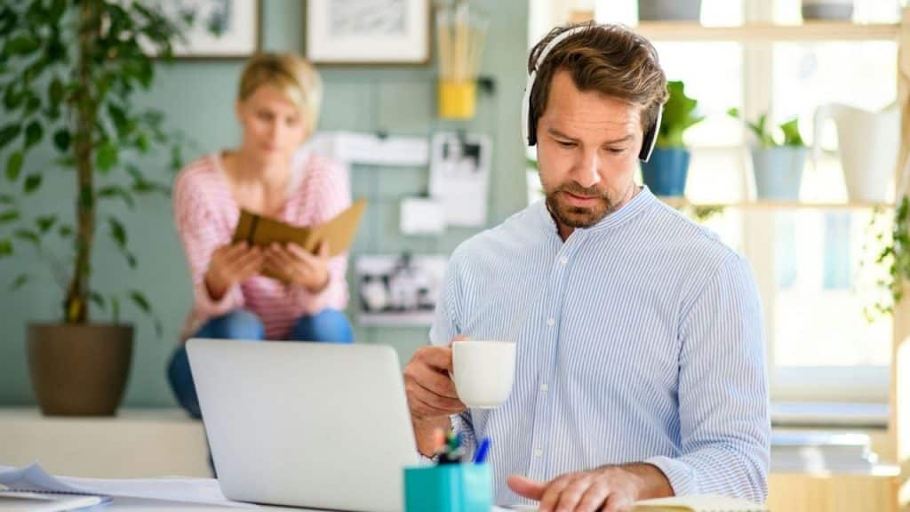 A man with headphones and coffee works at a laptop while a woman sits behind him.