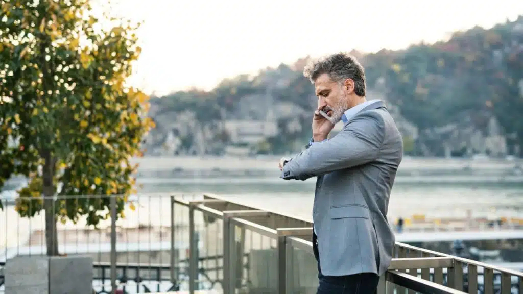 A man in a blazer checks his watch while talking on a cellphone outdoors near water.