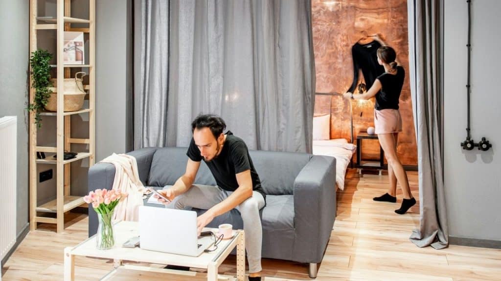 A man uses a laptop on a couch while a woman hangs clothes nearby.