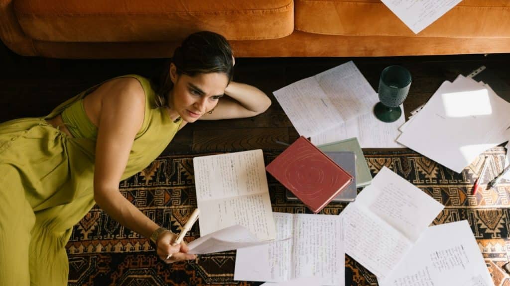 A woman in a green dress lies on a rug surrounded by many scattered papers.