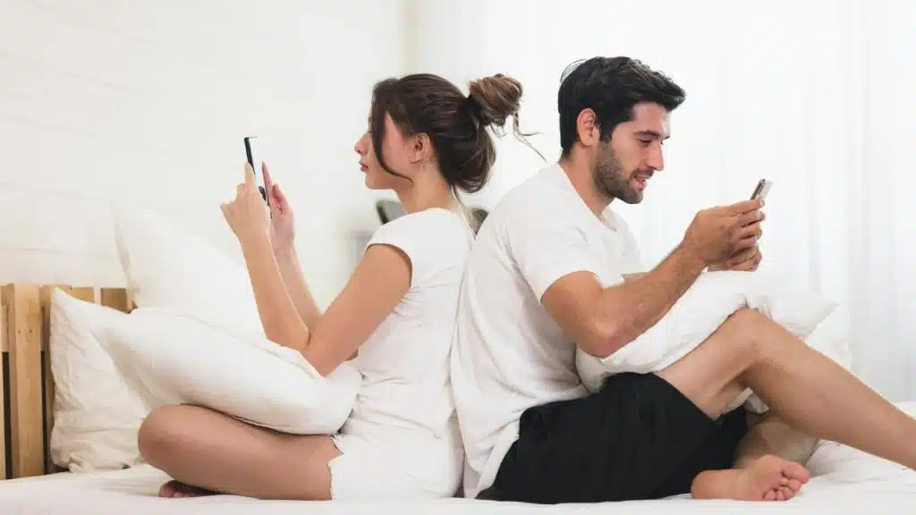 A man and woman sit back-to-back on a bed, each looking at their own smartphone.