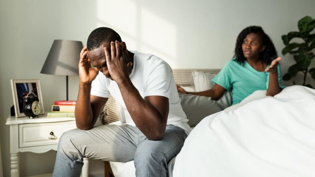 A man sits on a bed holding his head while a woman gestures in the background.