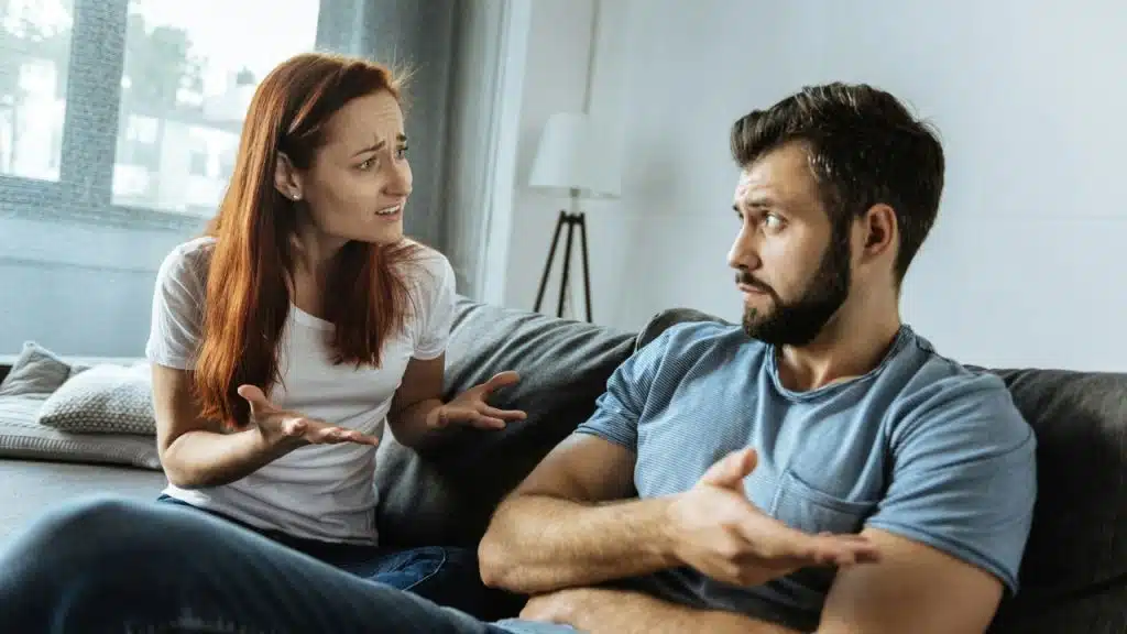 A woman with red hair gestures with her hands while talking to a bearded man.