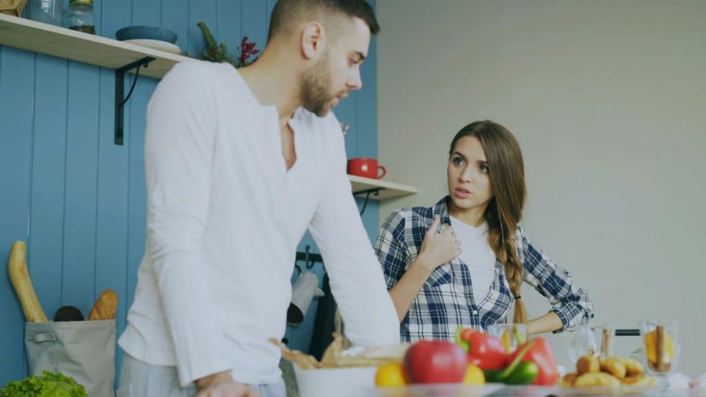 A woman in a plaid shirt speaks to a man standing in a kitchen setting.
