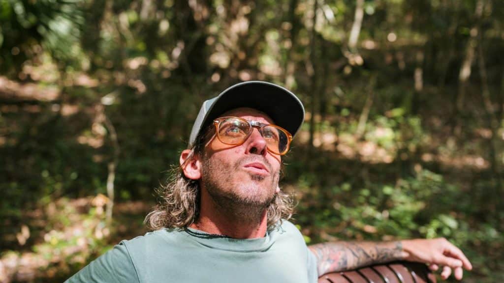 A man wearing glasses and a cap sitting on a bench in a wooded area.