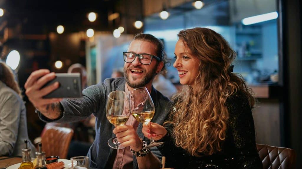 A couple smiling and taking a selfie while holding wine glasses at a restaurant.