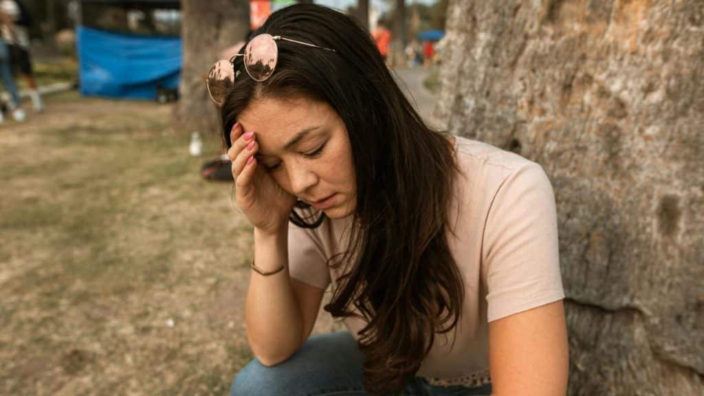 A woman with her hand on her forehead sits outdoors against the trunk of a tree.