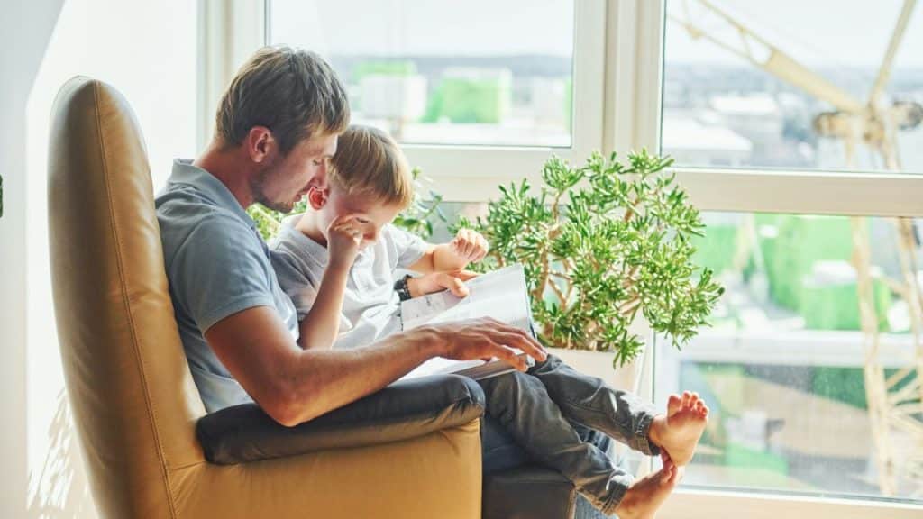 A man and a young child sit together in a large chair reading a book together.