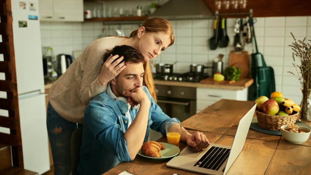 A woman stands behind a man seated at a laptop, resting her hand on his head.