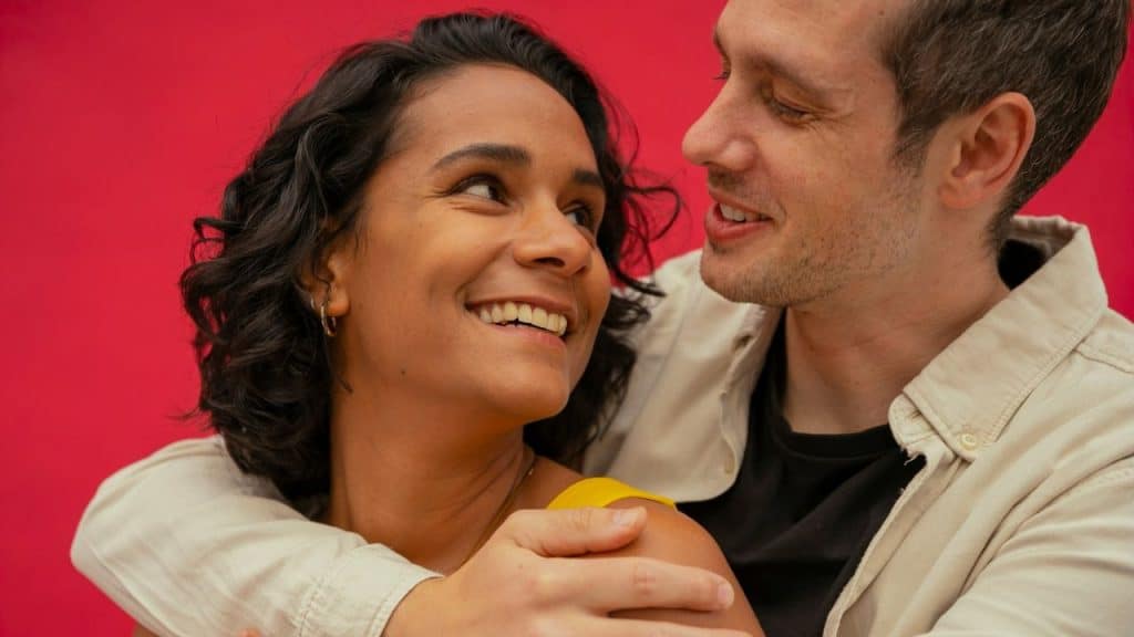 A man with light skin embraces a woman with dark curly hair against a red background.